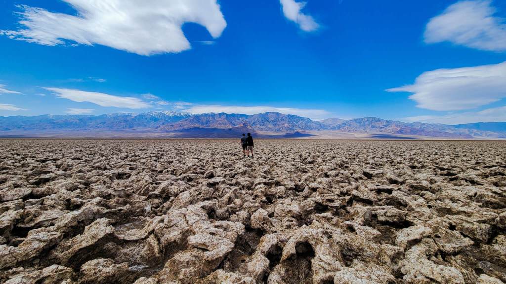 A boy and a teenager in black shirts and shorts stand next to each other in the distance looking away from the camera. In the background are3 mountains and a blue sky spotted with clouds. In the foreground are salt rock formations. Devil's Golf Course in Death Valley National Park, MPA Project Travels.