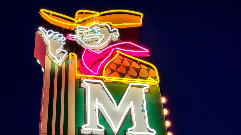 A neon sign against a dark sky. A cowboy smoking a cigarette with a thumb up sits on top of an M. tips for visiting the neon museum. MPA Project Travels.