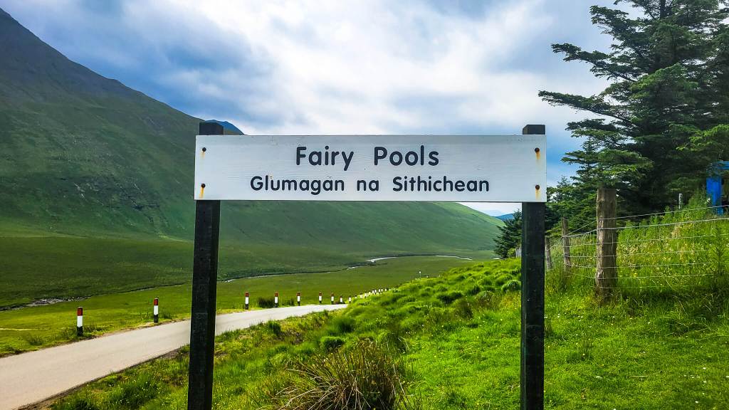 Fairy Pools trail head sign. A single lane road surrounded by green mountains.  Isle of Skye Fairy Pools MPA Project Travels
