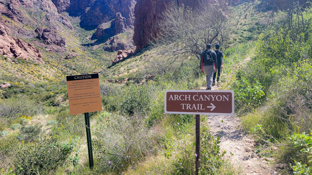 A man and a teenage boy walk away from the camera on a trail. In the foreground are two trail markers. One says Arch Canyon Trail in white letters on a brown background. The other is a yellow caution sign with small black writing. Arch Canyon Trail in Organ Pipe Cactus. MPA Project Travels