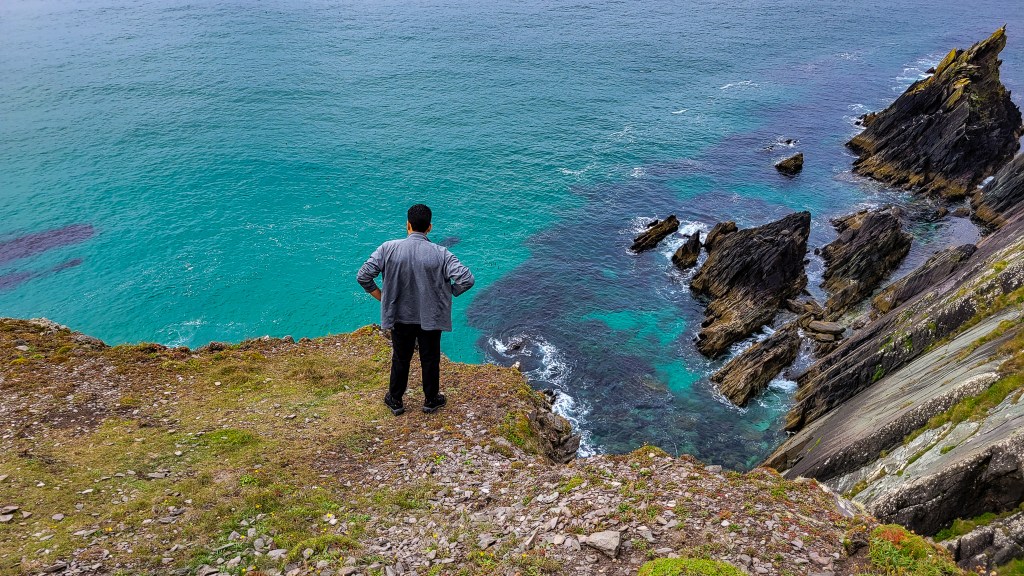 A man in black pants and a gray jacket looks out over a sea cliff onto turquoise water and the rocks below.